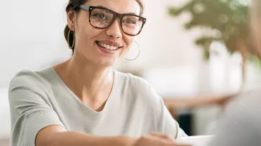Smiling woman with glasses sitting at a table during a conversation.