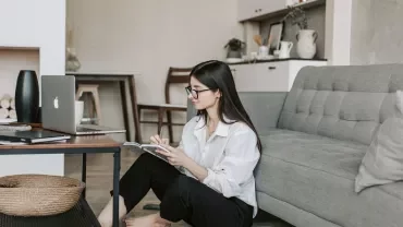 Woman sitting on floor near sofa, reading a book next to a laptop.