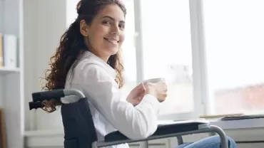 Woman in wheelchair smiling and holding a mug, sitting at a desk with a laptop.