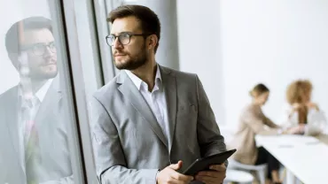 Businessman in a suit holding a tablet and looking out a window, with colleagues working at a table in the background.