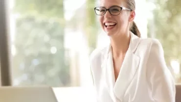 Smiling woman in glasses and a white blazer using a laptop.