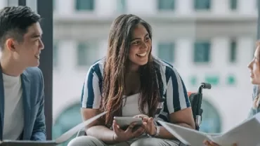 Three people in a meeting, smiling and discussing with papers and tablets.