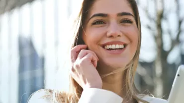 A Caucasian woman smiling with teeth while holding a mobile phone to her right ear, wearing a long-sleeved white top, in an outdoor setting with a building and some trees behind her.