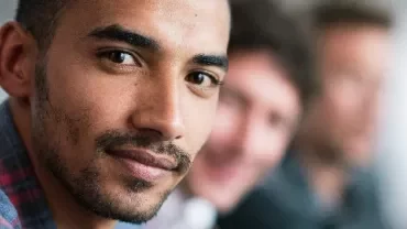 Man in a plaid shirt smiling with blurred colleagues in the background.