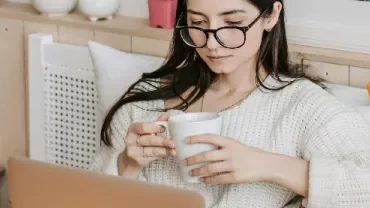 Woman with glasses sipping coffee while using a laptop.