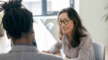 A woman in glasses converses with a man in an office setting, both engaged in discussion.