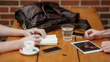 Two people at a table with coffee, notebook, tablet, glass of water, and a bag.