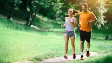 A man and woman jogging on a sunny path surrounded by green trees.