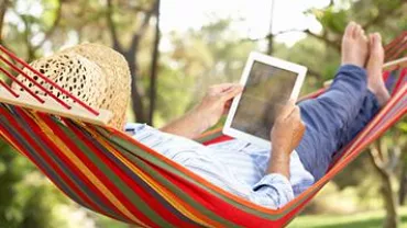 Person relaxing in a colorful hammock with a tablet on a sunny day.