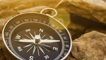 A compass resting on rocky ground, showing cardinal directions with sunlight casting a warm glow.