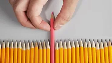 A hand picking a pink pencil out of a row of yellow pencils.