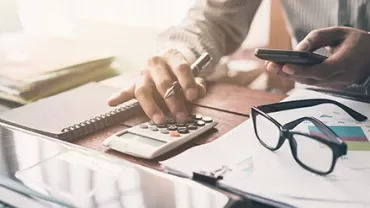 Person using a calculator at a desk with a smartphone, notepad, and glasses.
