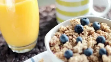 Bowl of granola with blueberries next to a glass of orange juice and a striped mug.