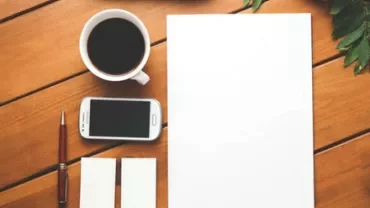 Workspace with coffee, smartphone, pen, and blank paper on a wooden table.