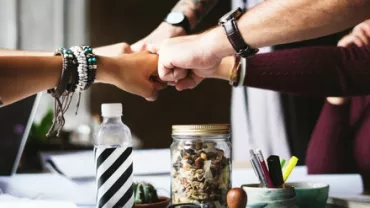 Four people fist bumping over a table with office supplies.