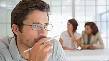Man thinking with pen in hand, two women in background.