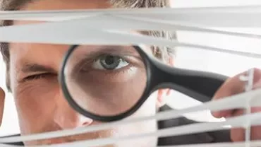 Person looking through blinds with a magnifying glass.