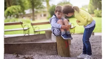 Child sitting on a wooden post while another child leans in, in a park setting.