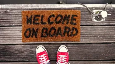 Doormat on wooden dock reads 'Welcome On Board' with red sneakers visible.