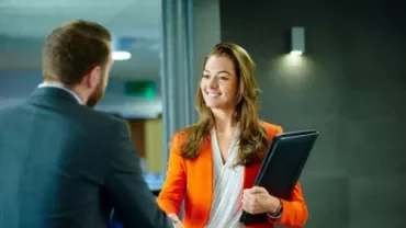 Woman in orange blazer shakes hands, holding a folder.
