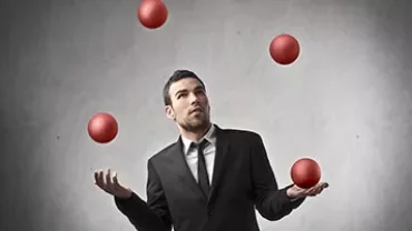 Man in a suit juggling four red balls with a focused expression.