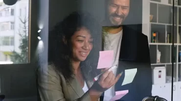 Two colleagues smiling while brainstorming with sticky notes on glass.