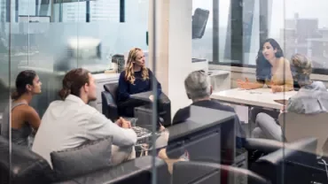 Business meeting in glass-walled office with people discussing around a table.