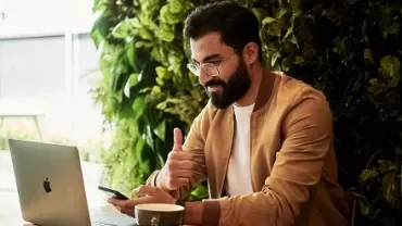 Man giving thumbs up while using laptop and phone in a cafe.