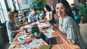 Woman smiling at coworking space with laptop and phone.