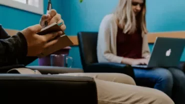 Person taking notes while another works on a laptop in a waiting room.
