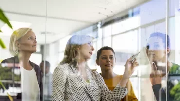 Business meeting with four women discussing in front of a glass board.