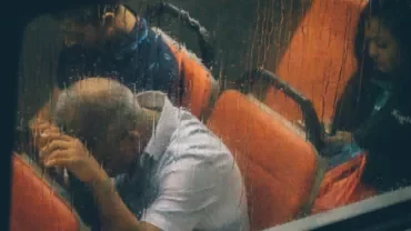 Three people sitting on a bus in the rain, viewed through a wet window.