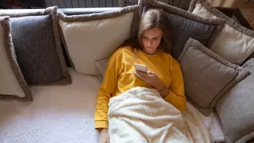 Person in yellow shirt lying on a sofa with pillows, looking at a phone.