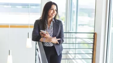 Woman standing in a modern office, holding a phone and smiling.