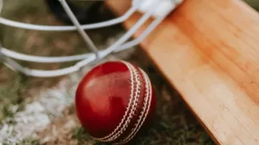 Close-up of a red cricket ball, a wooden bat, and a helmet on grass.