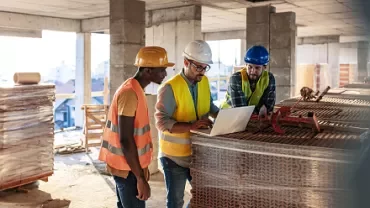 Three construction workers in safety gear reviewing plans on a laptop.