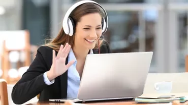 Smiling woman with headphones waves during a video call on a laptop.
