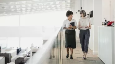Two women walking and talking in a modern office hallway.