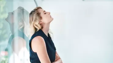 Woman in blue top leaning against glass wall, eyes closed.