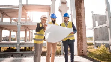 Three construction workers in safety vests and helmets review blueprints at a building site.