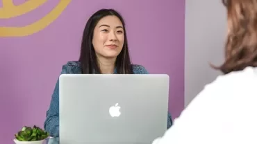 Woman sitting at a table using a laptop, smiling across from another person.