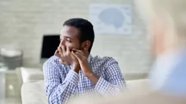 Man in a checkered shirt looking pensive in a bright room.