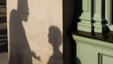 Silhouettes of two people in conversation cast on a wall beside a green building column.