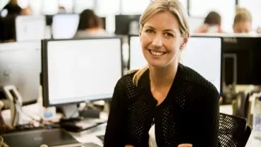Smiling woman at office desk with computer monitors in the background.