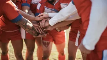Rugby team huddles with hands together before a match, wearing red uniforms.