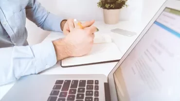 Person writing in a notebook at a desk with a laptop and plant.