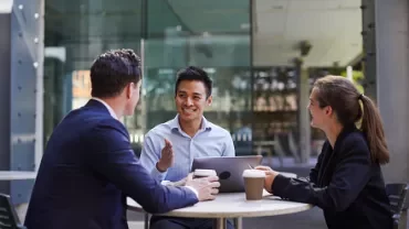 Three people having a meeting at an outdoor cafÃ© table, smiling with coffee cups and a laptop.