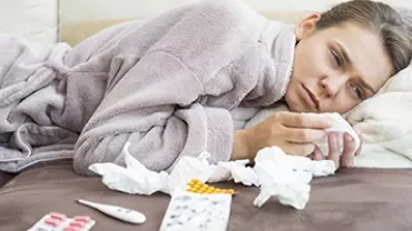 Woman in bathrobe lying on a couch surrounded by tissues and medicine packs.