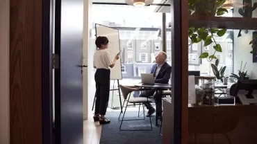 A woman stands at a whiteboard presenting to a seated man in an office with large windows.