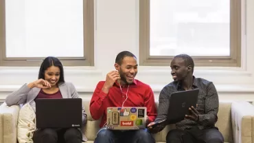 Three people sitting on a couch using laptops, smiling and talking to each other.